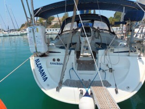 The stern of a sailboat named "ALEMANIA" in the harbor, equipped with a liferaft and a steering wheel, connected to the dock by a ramp.
