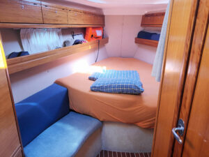 Eine Kabine auf einem Segelboot mit einem großen Bett in der Mitte, orangefarbener Bettwäsche, blauen Kissen und Stauraum an den Seitenwänden.